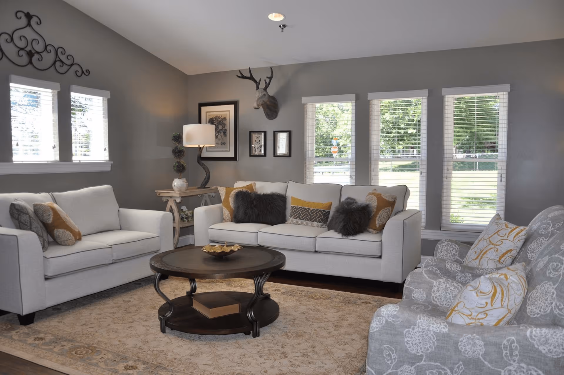 A cozy living room with two light gray sofas and a patterned armchair arranged around a dark wooden round coffee table. The room has gray walls, several windows with white blinds letting in natural light, a floor lamp, framed artwork, and a decorative mounted deer head on the wall.