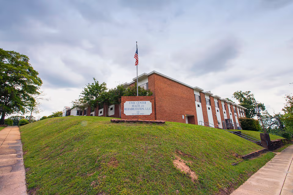 Exterior view of Civic Center Health and Rehabilitation, LLC, a two-story brick building situated on a grassy hill with a sidewalk leading up to it. An American flag flies on a flagpole near the building's sign.