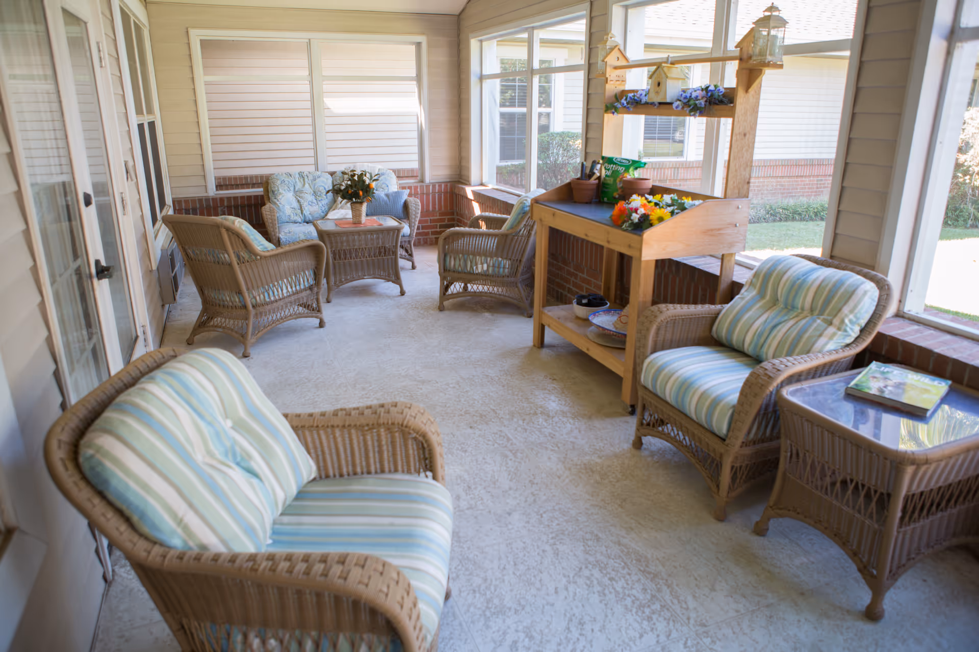 A bright enclosed porch area with wicker furniture including armchairs with striped cushions, a loveseat with floral cushions, and a small table with a flower vase. There is a wooden potting bench with gardening supplies and decorative birdhouses. Large windows provide a view of the outside garden area.