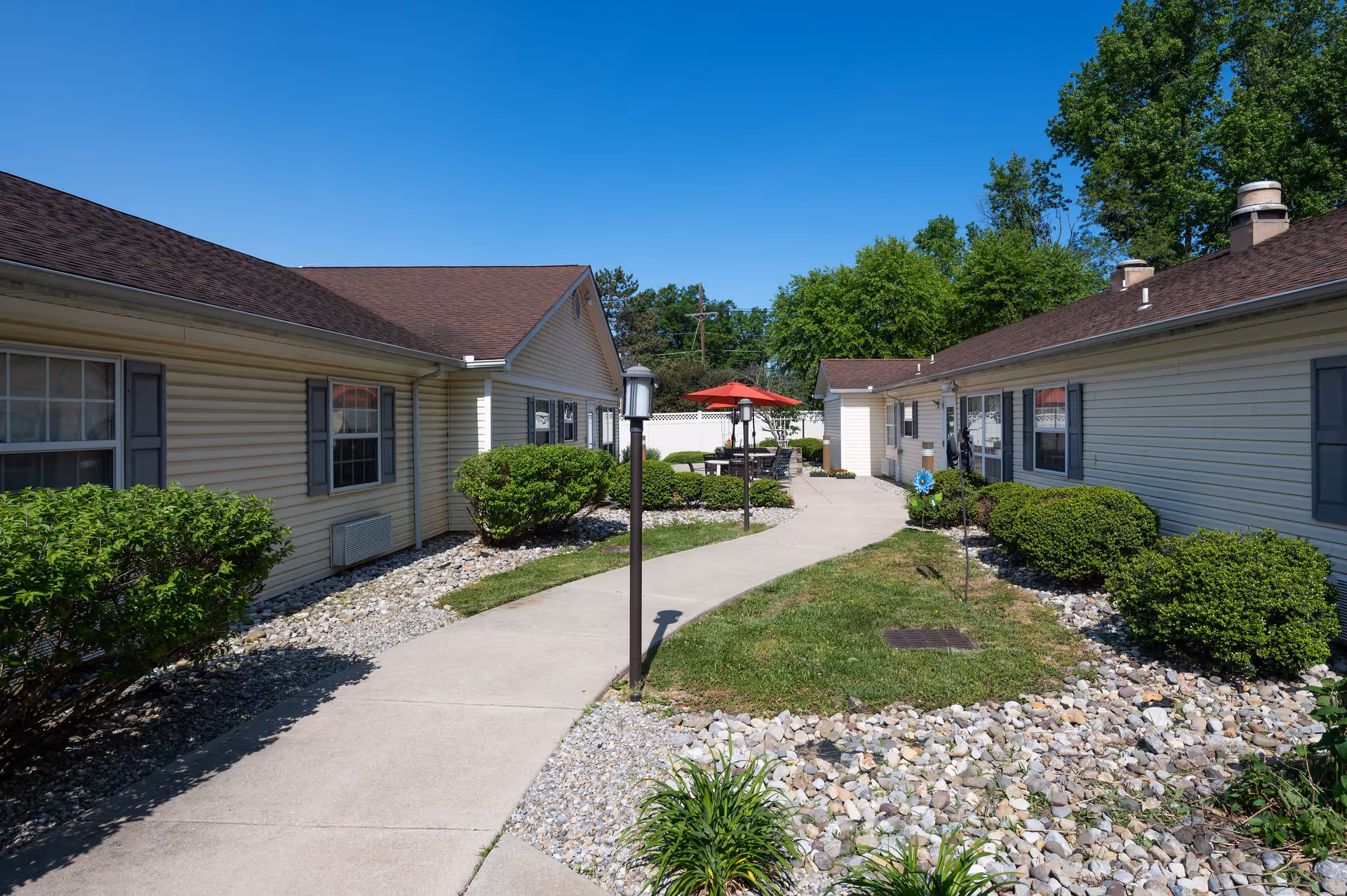 Outdoor view of a senior living facility courtyard with a concrete walkway winding between two single-story buildings with beige siding and brown roofs. The area is landscaped with bushes, rocks, and small patches of grass. There is a seating area with a table and red umbrella in the background under a clear blue sky.