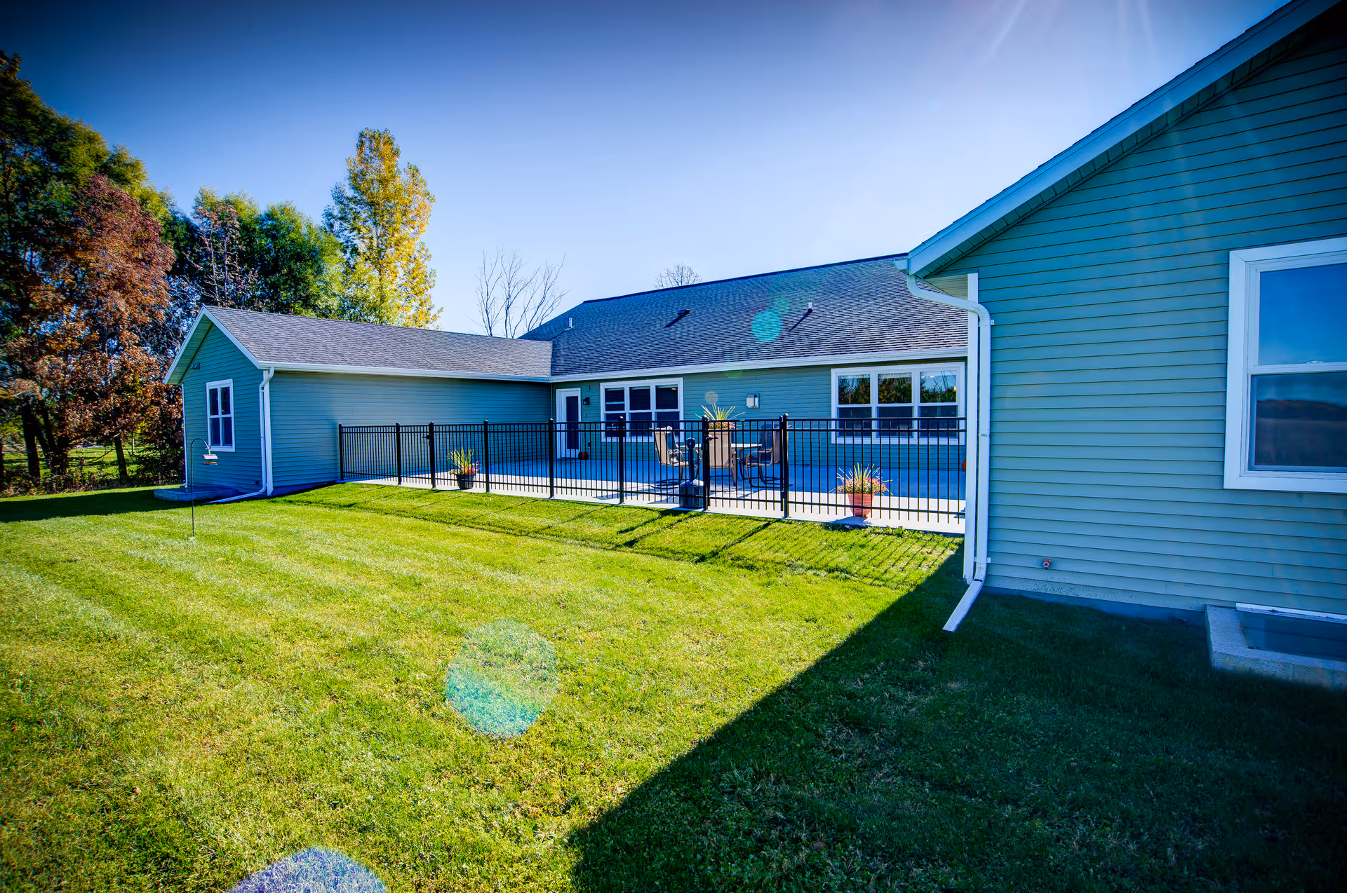 Exterior view of a single-story building with light blue siding and a fenced patio area with outdoor seating, surrounded by a well-maintained green lawn and trees under a clear blue sky.