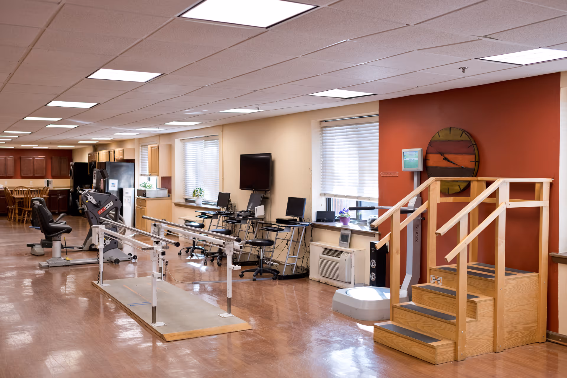 A spacious rehabilitation room with exercise equipment including parallel bars for walking practice, a stationary exercise bike, and a set of wooden stairs with handrails. The room has large windows with blinds, several computer workstations, and a wall clock on a red accent wall. In the background, there is a kitchen area with wooden cabinets and a dining table with chairs.