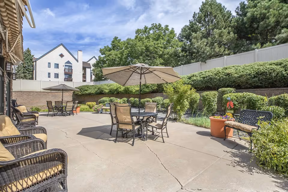 Outdoor patio area with several tables and chairs, some shaded by large umbrellas. The patio is surrounded by greenery including bushes, trees, and potted plants. A white fence and a building with white siding and dark trim are visible in the background under a partly cloudy sky.
