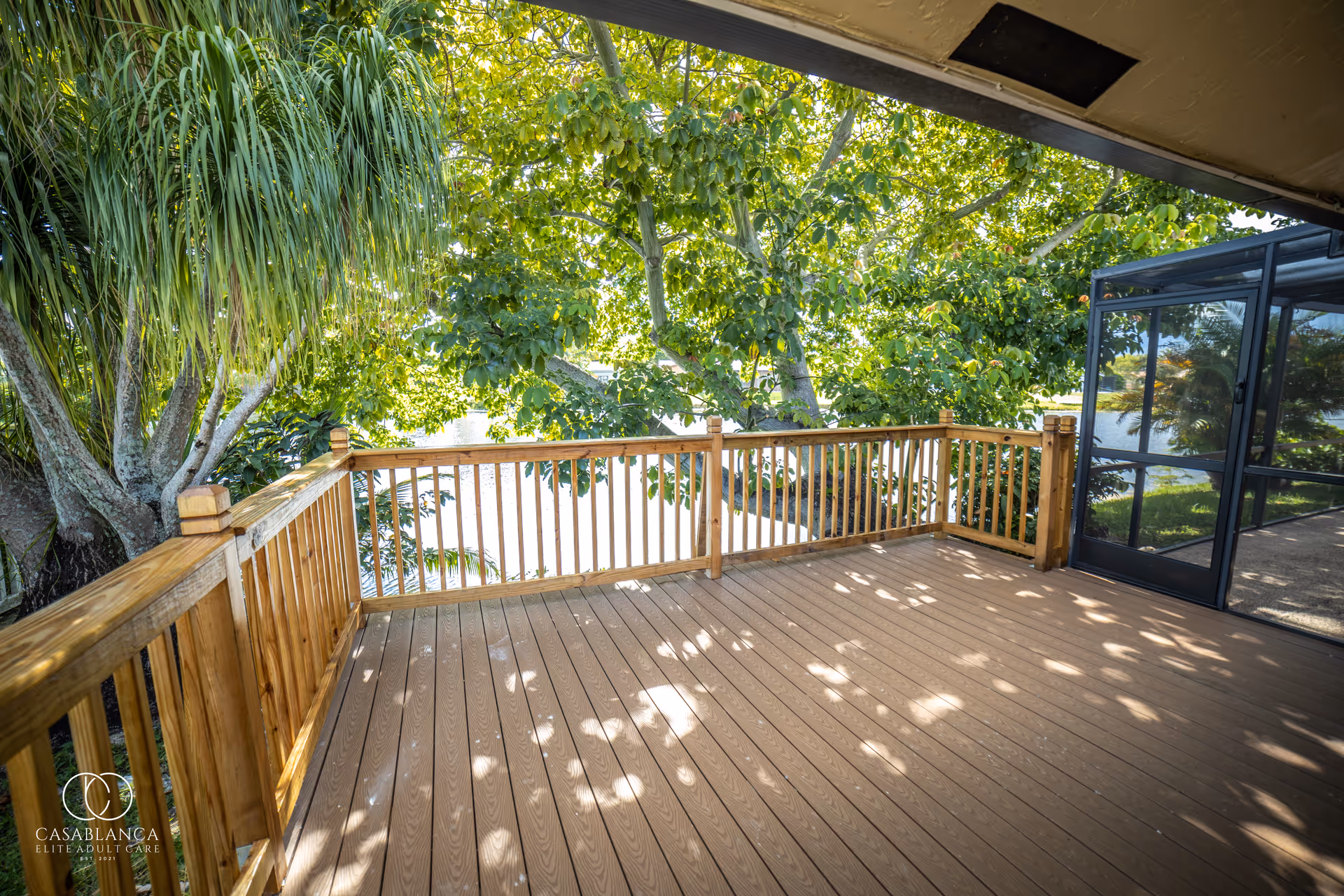 A wooden deck with railing overlooking a lush green tree and a body of water. The deck is partially shaded and adjacent to a screened-in porch area.