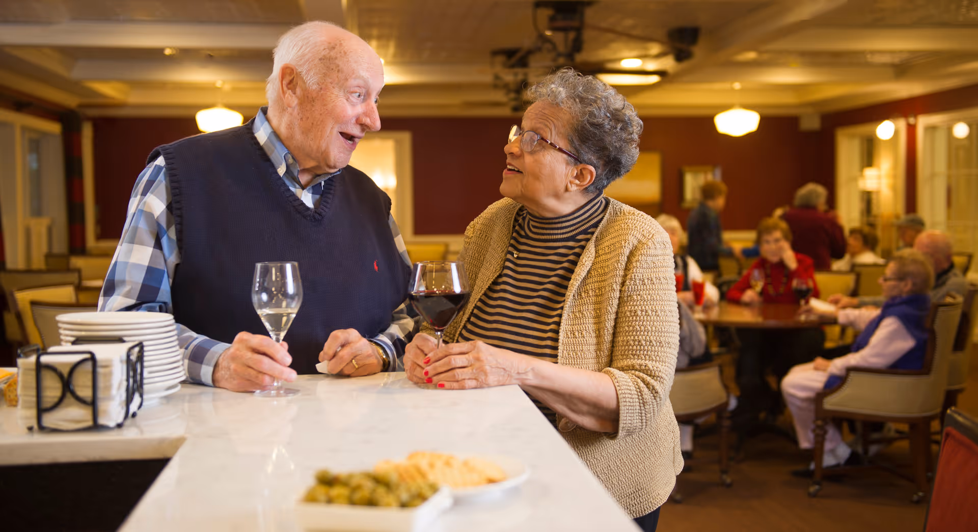 An elderly man and woman are standing at a counter in a warmly lit dining or common area, holding glasses of wine and engaging in a lively conversation. In the background, several other elderly people are seated at tables, socializing and enjoying the space.
