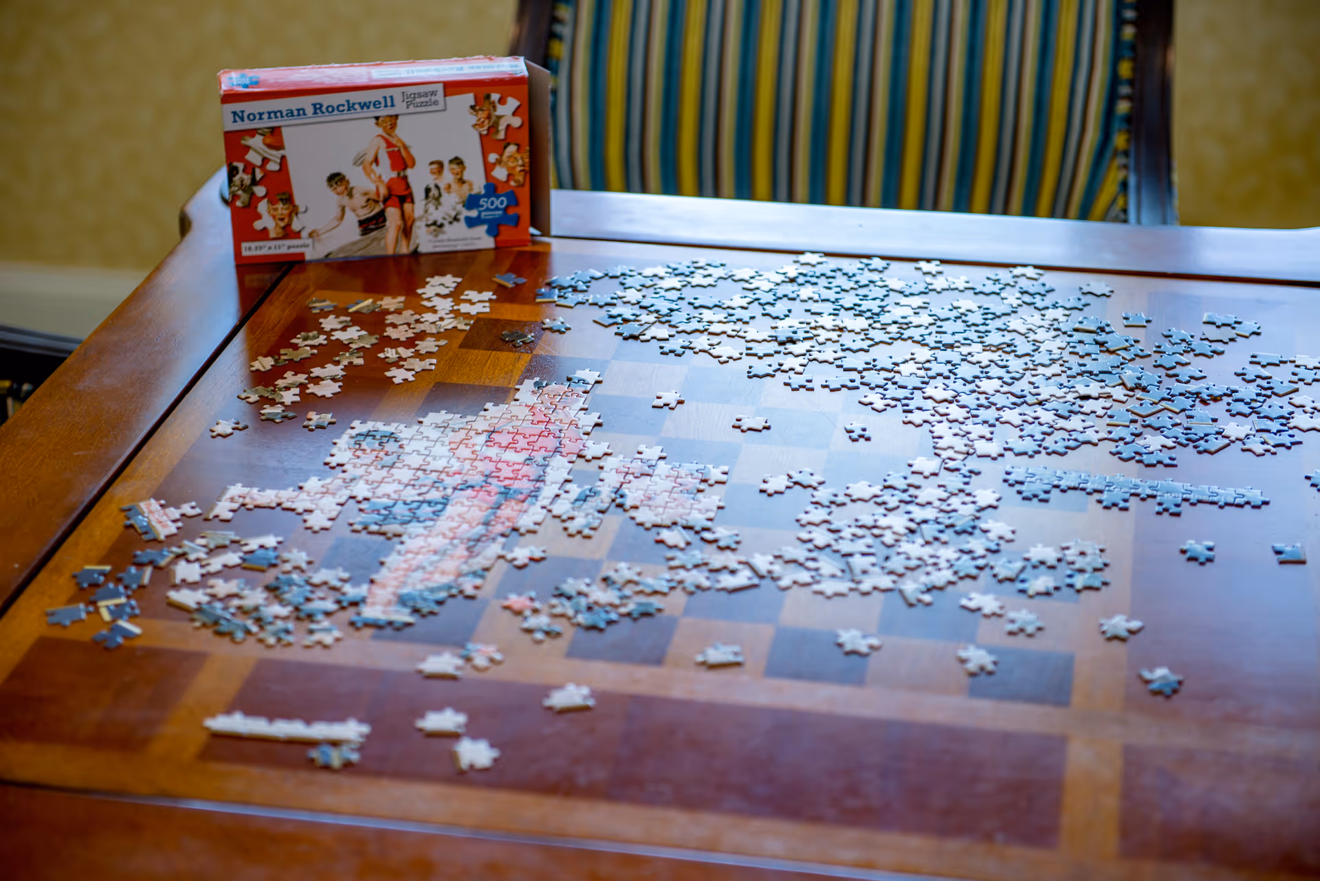 A wooden table covered with scattered jigsaw puzzle pieces and a Norman Rockwell puzzle box, with a striped chair behind it.