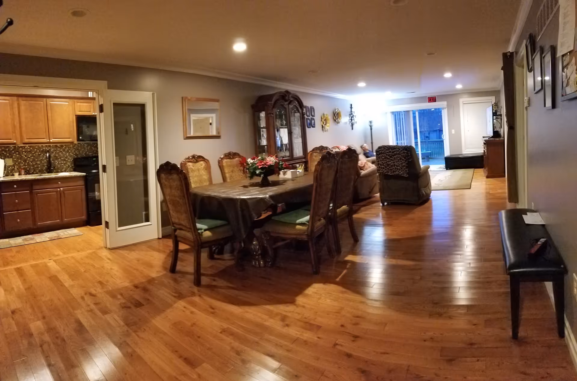 Open-plan dining and living area with a long dining table and chairs, hardwood floors, a kitchen to the left, and a seating area toward the back.