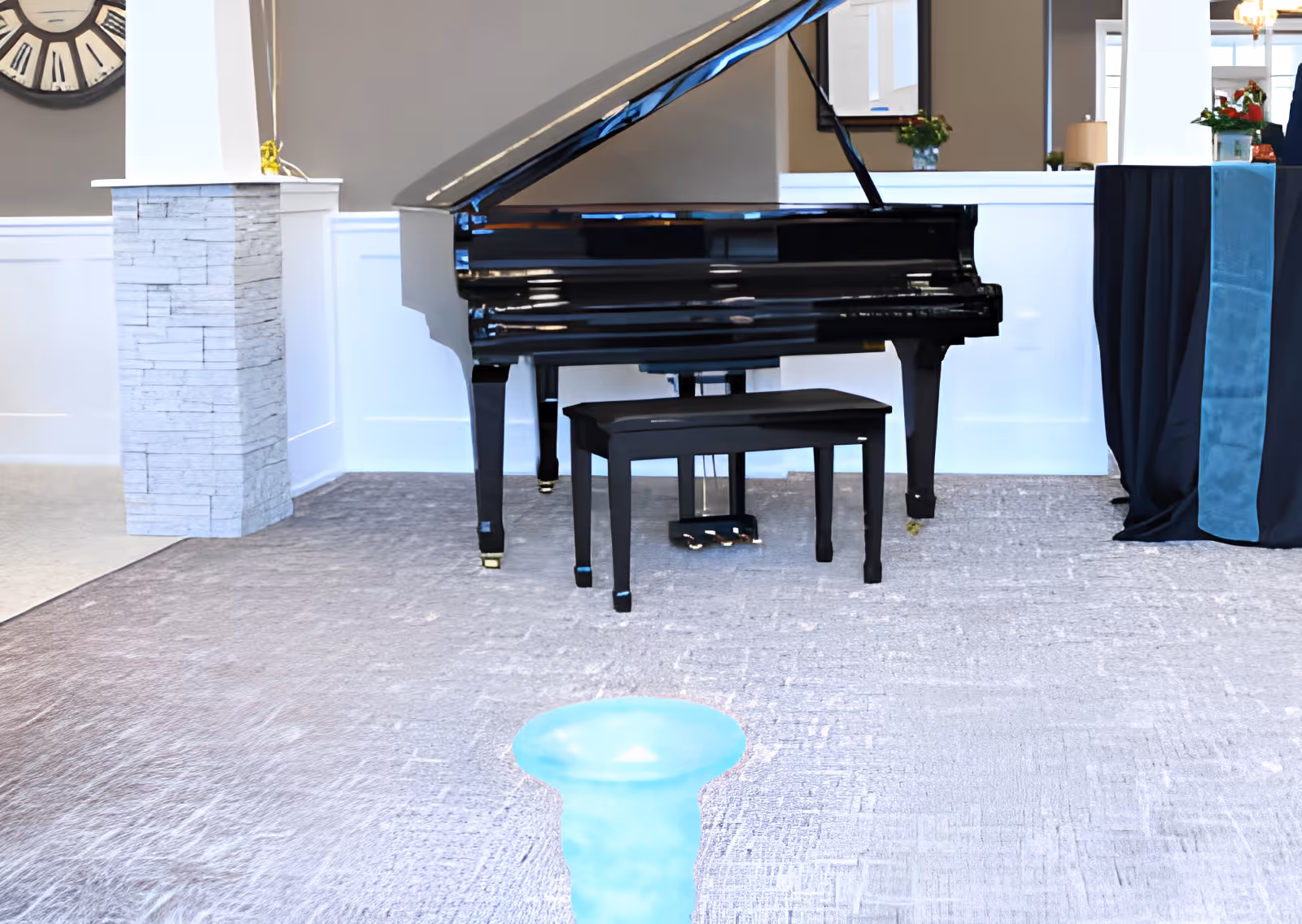 A black grand piano with a matching bench is placed on a carpeted floor in a well-lit room. The background features white wainscoting on the walls, a large decorative clock, a stone pillar, and a table covered with a black and blue cloth. There are also some plants and a mirror on the wall behind the piano.