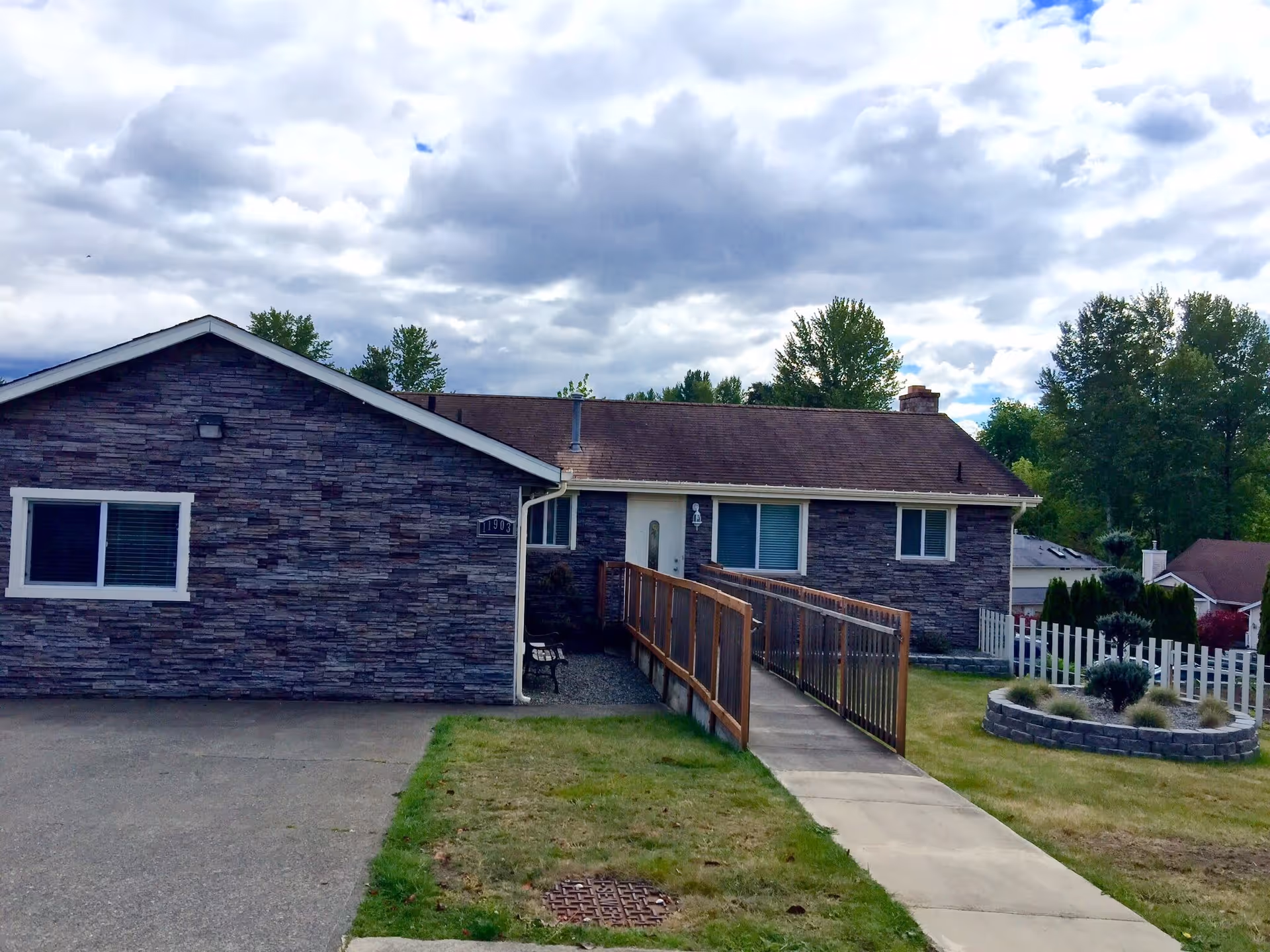 Exterior view of a single-story adult family home with stone facade, a sloped roof, and a wooden ramp leading to the front door. The house is surrounded by a lawn with a circular raised garden bed and trees in the background under a cloudy sky.