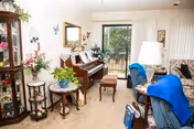 A cozy living room with a piano against the wall, a small round table with plants, a floral sofa, a blue armchair with a blue blanket, a floor lamp, and a sliding glass door leading to a balcony or patio.