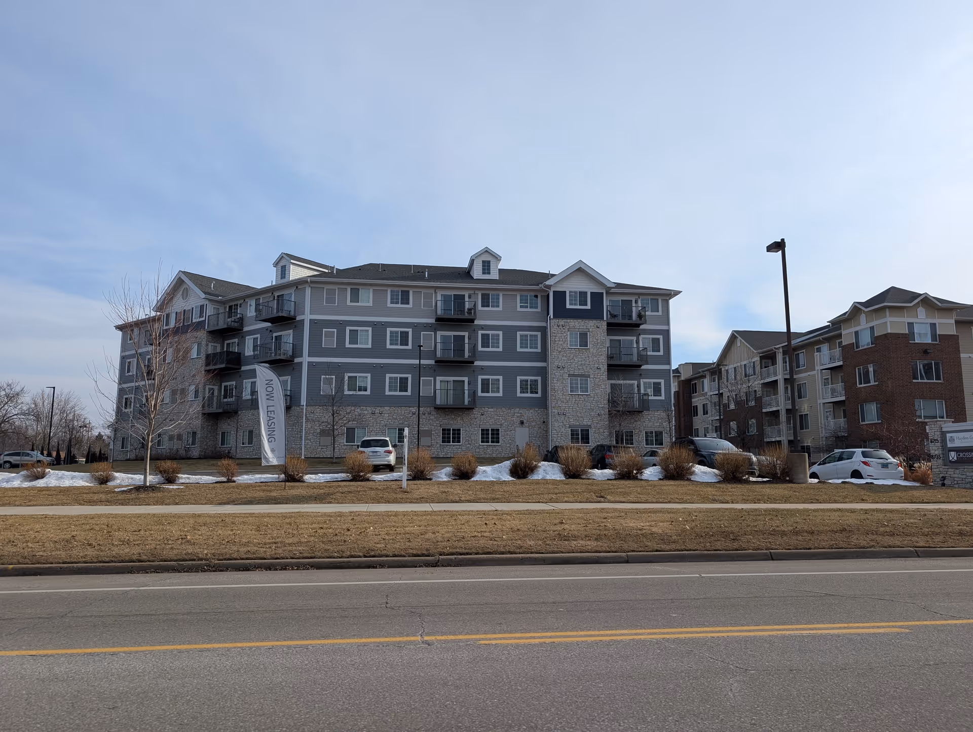 Exterior view of a multi-story senior housing building named Hayden Grove Senior Housing with balconies, parked cars, and a 'Now Leasing' flag on the lawn. The sky is clear with some clouds.