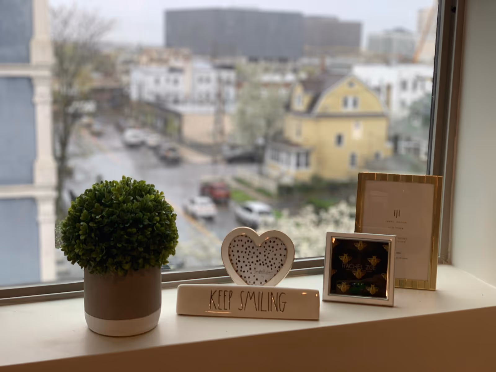 A windowsill with a small potted green plant, a heart-shaped decorative item, a sign that says 'KEEP SMILING', and two framed pictures. Outside the window, a blurred view of a street with cars and buildings is visible.