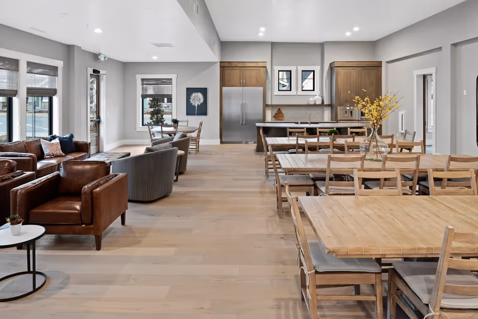 Spacious communal dining and lounge area with wooden tables and chairs, leather armchairs, and a kitchen in the background.