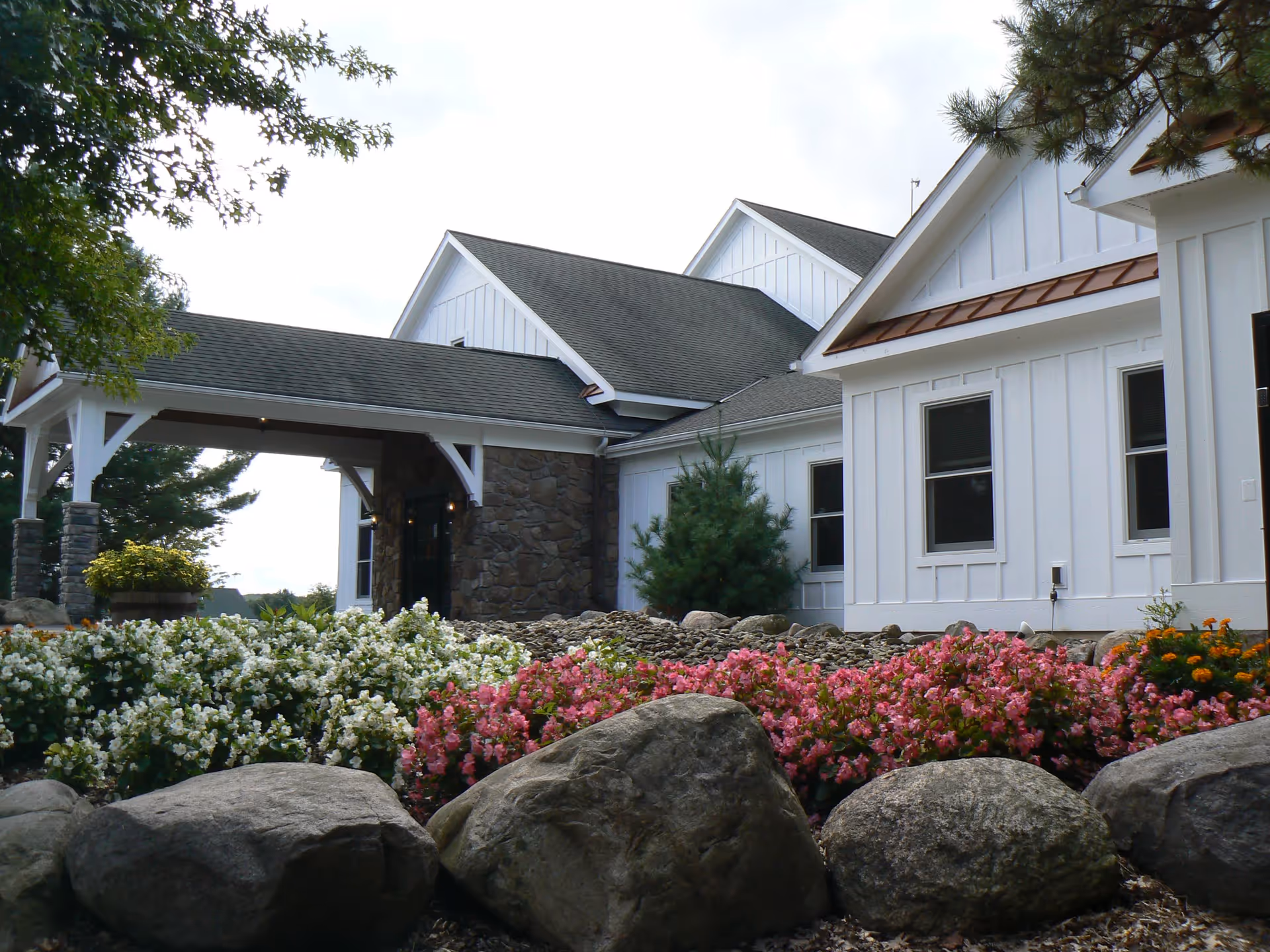 Exterior view of a white building with a stone entrance and a covered porch, surrounded by landscaped flower beds with pink and white flowers and large rocks in the foreground.