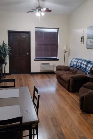 A cozy living room with wooden flooring, a brown loveseat with a blue and white checkered blanket draped over the back, a matching brown armchair, a floor lamp, a window with closed blinds, a ceiling fan with lights, a dark wooden door, and a dining table with chairs in the foreground.