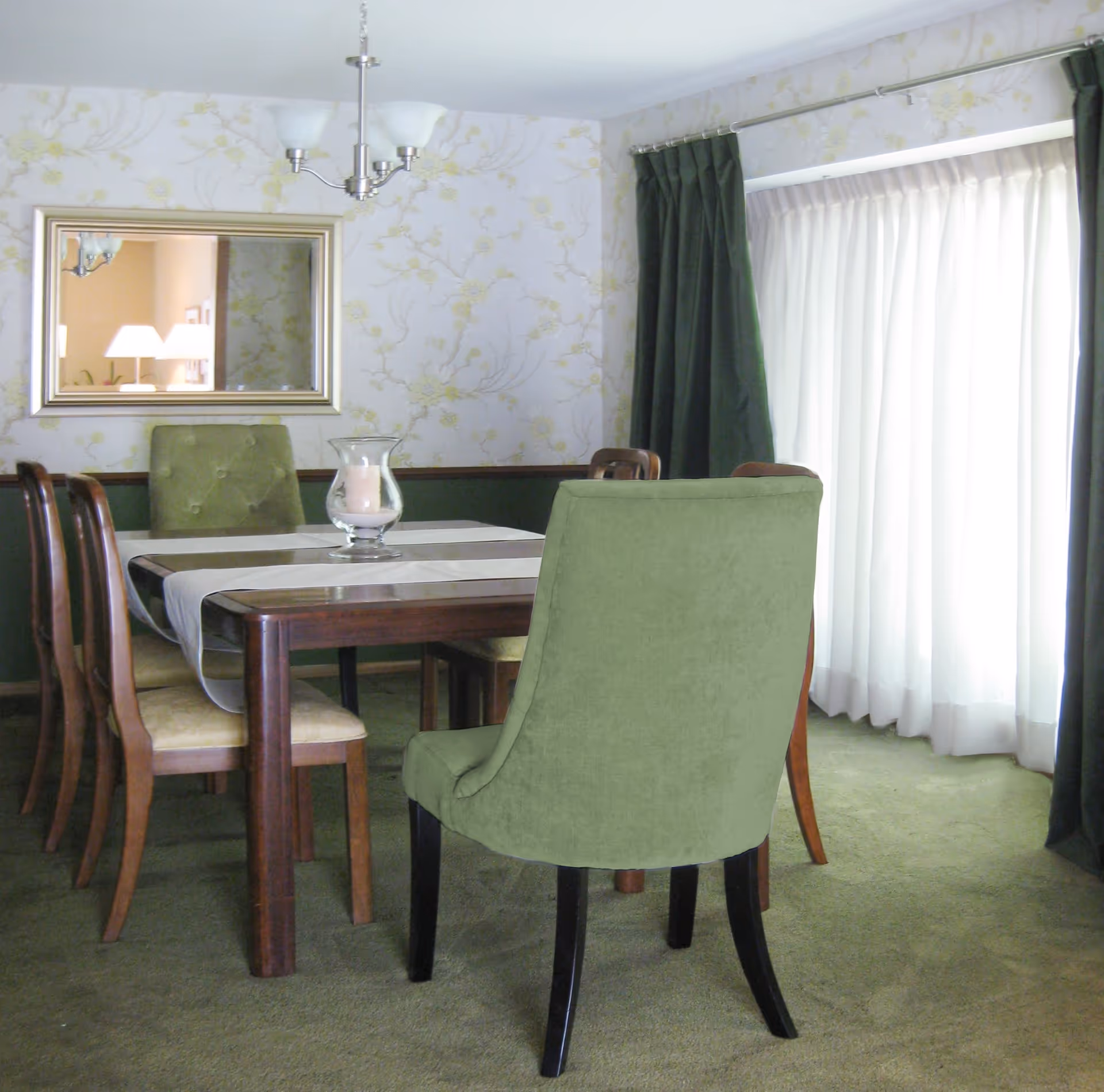 A dining room with a wooden table covered by a white table runner and a glass candle holder centerpiece. There are six chairs around the table, with two upholstered green chairs at the ends and four wooden chairs with beige cushions on the sides. The room has green carpet, floral wallpaper, a large mirror on the wall, and a window with sheer white curtains and dark green drapes.
