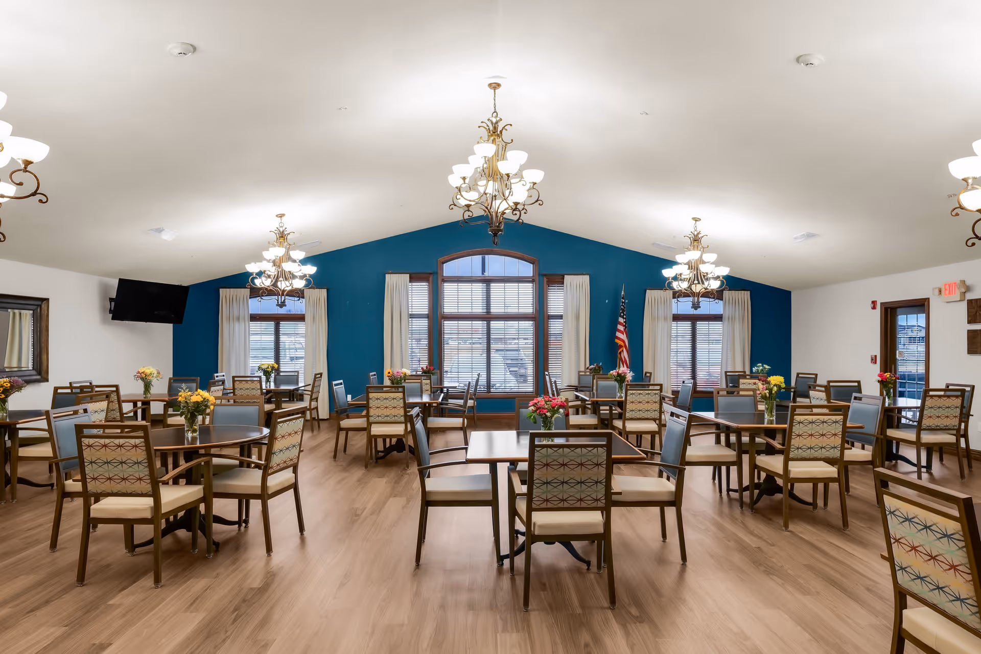 Spacious dining room with multiple tables and chairs, chandeliers, large windows, and floral centerpieces.