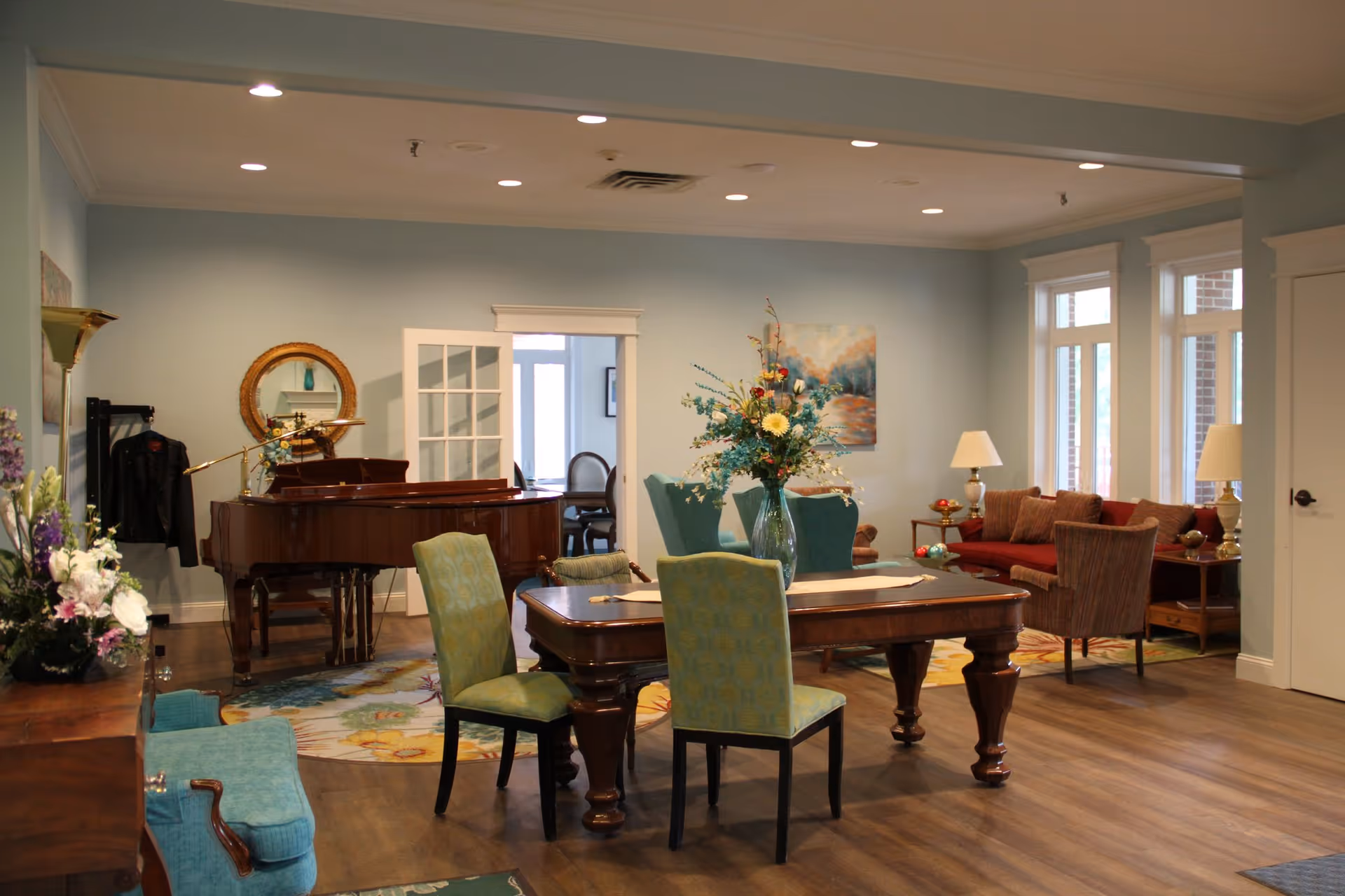 A cozy living room area in Illinois Presbyterian Home Communities featuring a wooden dining table with four green upholstered chairs, a grand piano in the corner, a floral centerpiece on the table, a red sofa with matching armchairs, two table lamps, and large windows letting in natural light.