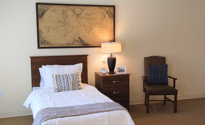 Single-occupancy bedroom with a made bed, nightstand holding a lamp and alarm clock, framed world map above, and a chair to the right.