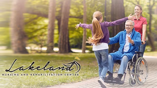An elderly man in a wheelchair is outdoors on a paved path in a park-like setting, smiling and reaching out to a young girl with long hair who is running towards him with open arms. A woman stands behind the wheelchair, smiling. Trees and greenery are visible in the background.
