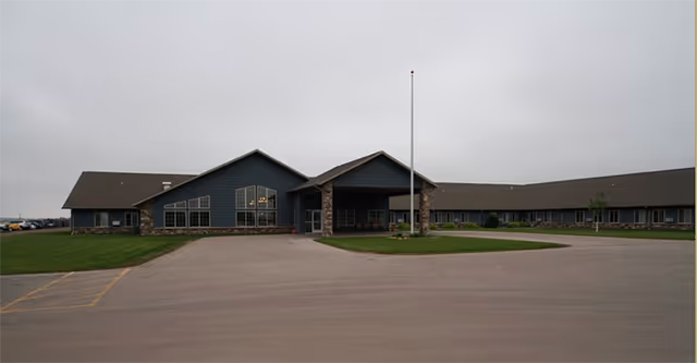 Exterior view of a single-story assisted living facility building with a large parking area in front, a flagpole near the entrance, and a cloudy sky overhead.