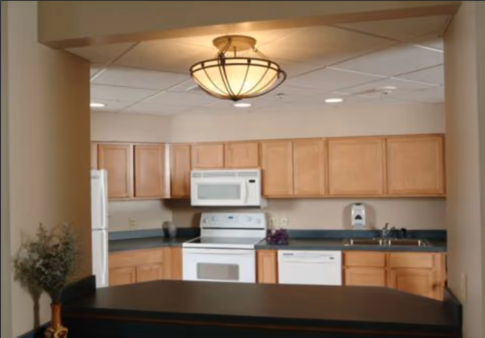 Open kitchen with light wood cabinets, white appliances, a dark countertop island and an overhead light fixture.