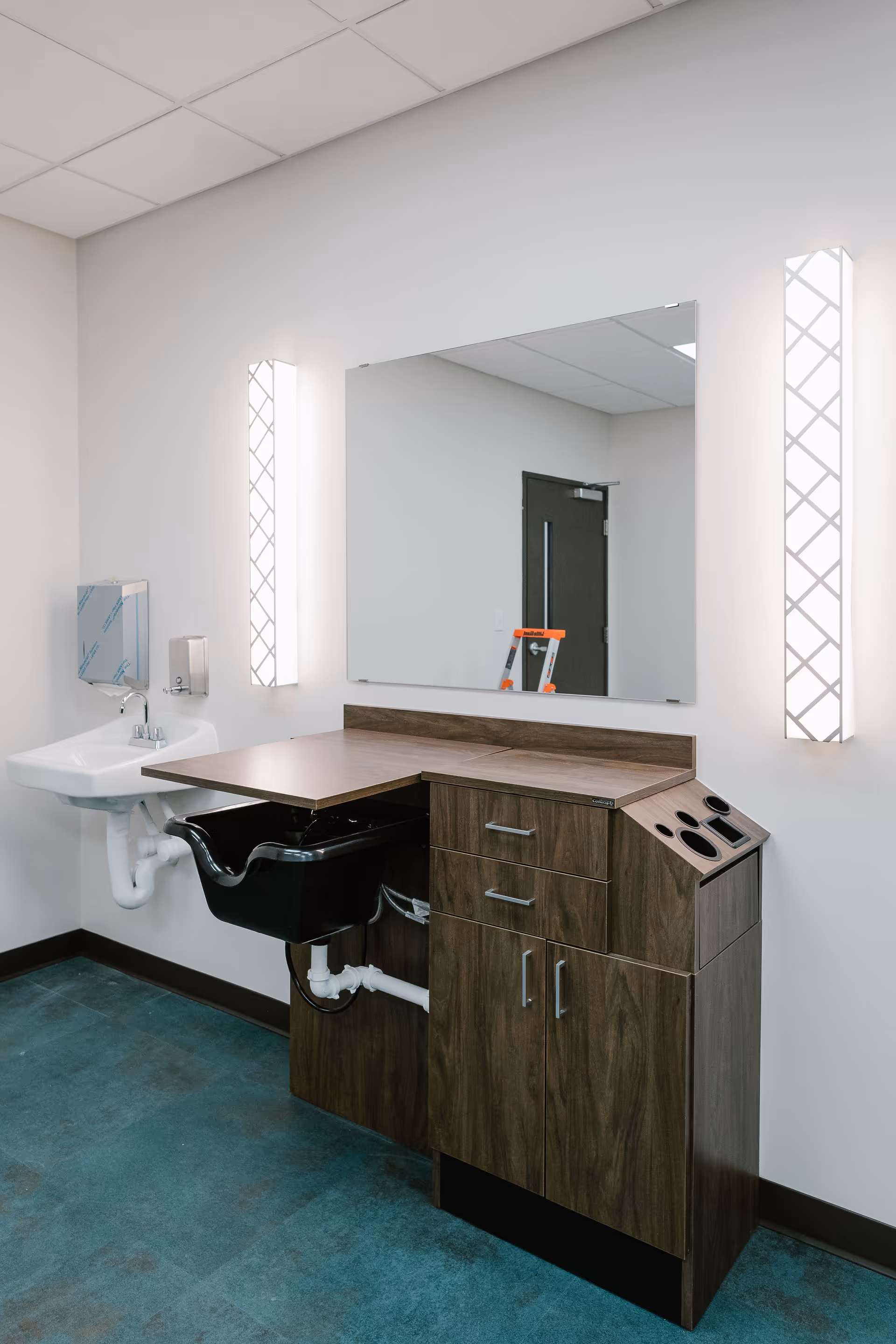 A clean, modern grooming station with a large rectangular mirror mounted on the wall, flanked by two vertical light fixtures. Below the mirror is a wooden cabinet with drawers and compartments, including a built-in black basin. To the left is a white wall-mounted sink with a soap dispenser and paper towel holder above it. The room has a blue floor and white walls with a dark door reflected in the mirror.