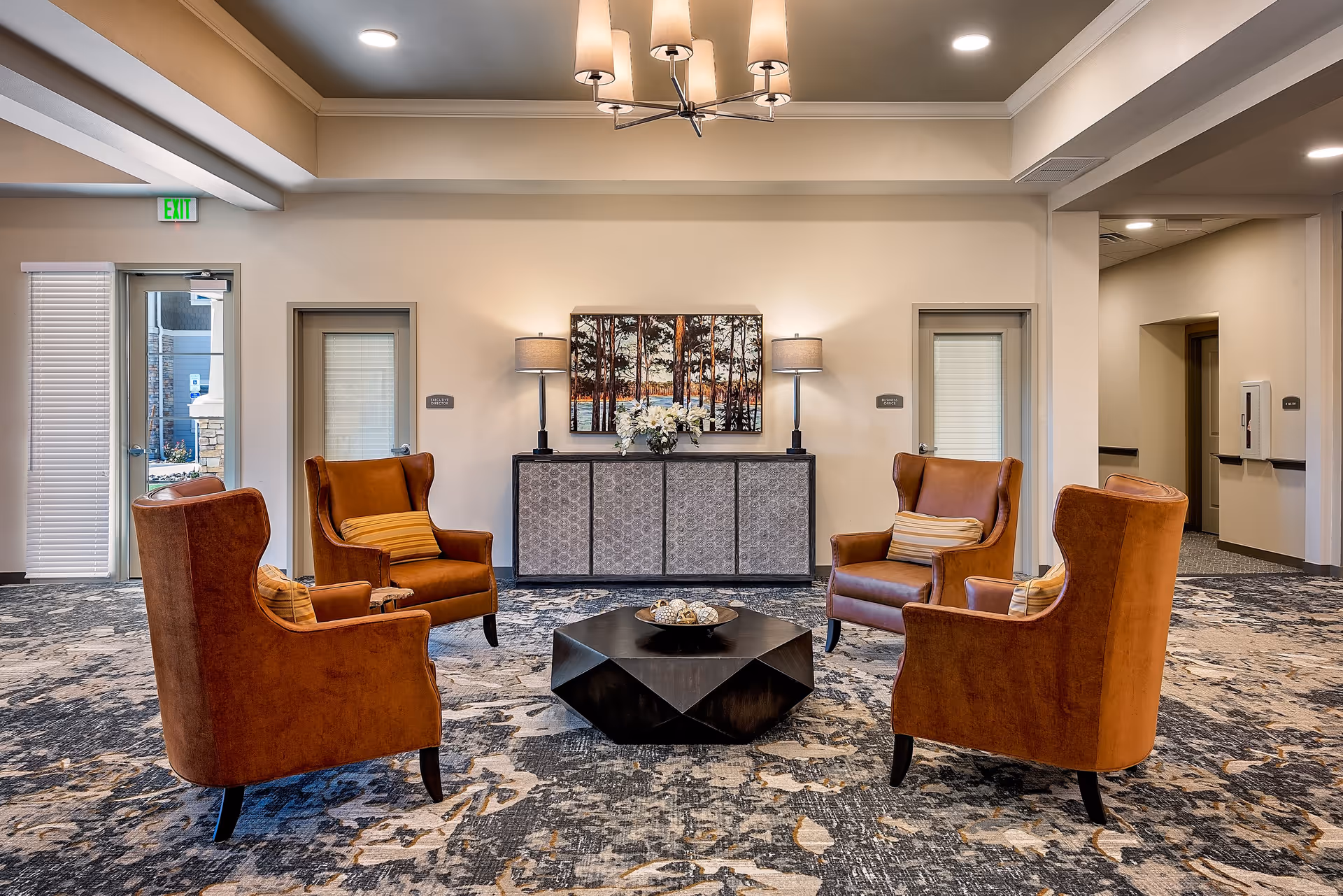 A cozy seating area in a senior living facility with four brown leather armchairs arranged around a black geometric coffee table. Behind the chairs is a sideboard with two table lamps and a vase of white flowers, and a painting of trees on the wall. The room has a patterned carpet and neutral-colored walls with recessed lighting.