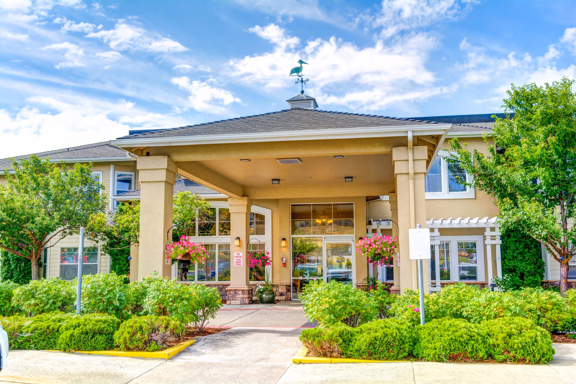 Front entrance of a senior living facility with a covered drop-off area, surrounded by green bushes and trees, hanging flower baskets, and a blue sky with some clouds above.