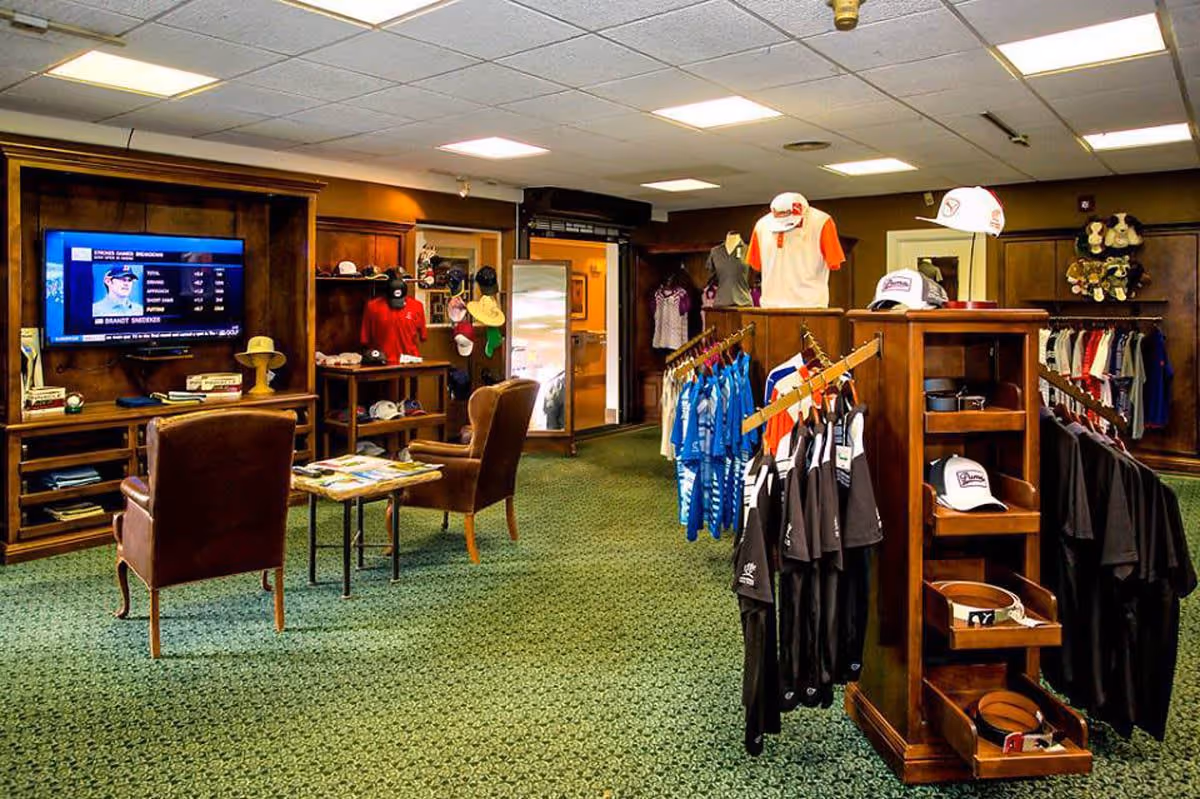 Interior view of a retail shop area within a senior living facility featuring wooden shelves and racks displaying clothing, hats, and accessories. Two brown leather chairs face a wall-mounted TV showing a sports broadcast. The room has green patterned carpet and a drop ceiling with fluorescent lights.