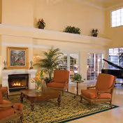 Bright communal lounge with upholstered armchairs and a coffee table arranged on a patterned rug, a lit fireplace, potted plants, and a grand piano under high ceilings.