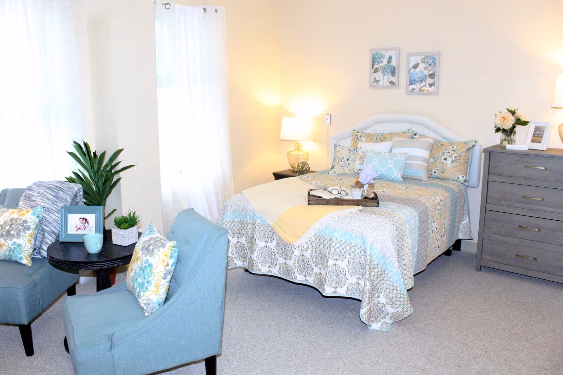 Sunlit furnished bedroom with a made bed, two blue armchairs, a dresser, lamps, and decorative plants and pillows.