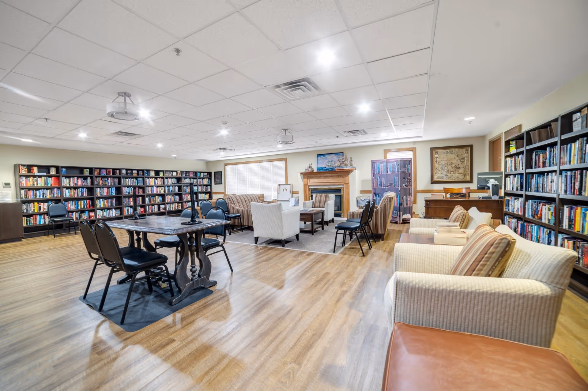 A spacious and well-lit library room with wooden floors, bookshelves filled with books along the walls, a wooden table with chairs in the center, and comfortable armchairs and sofas arranged around a fireplace. The room has a cozy and inviting atmosphere with framed artwork on the walls.