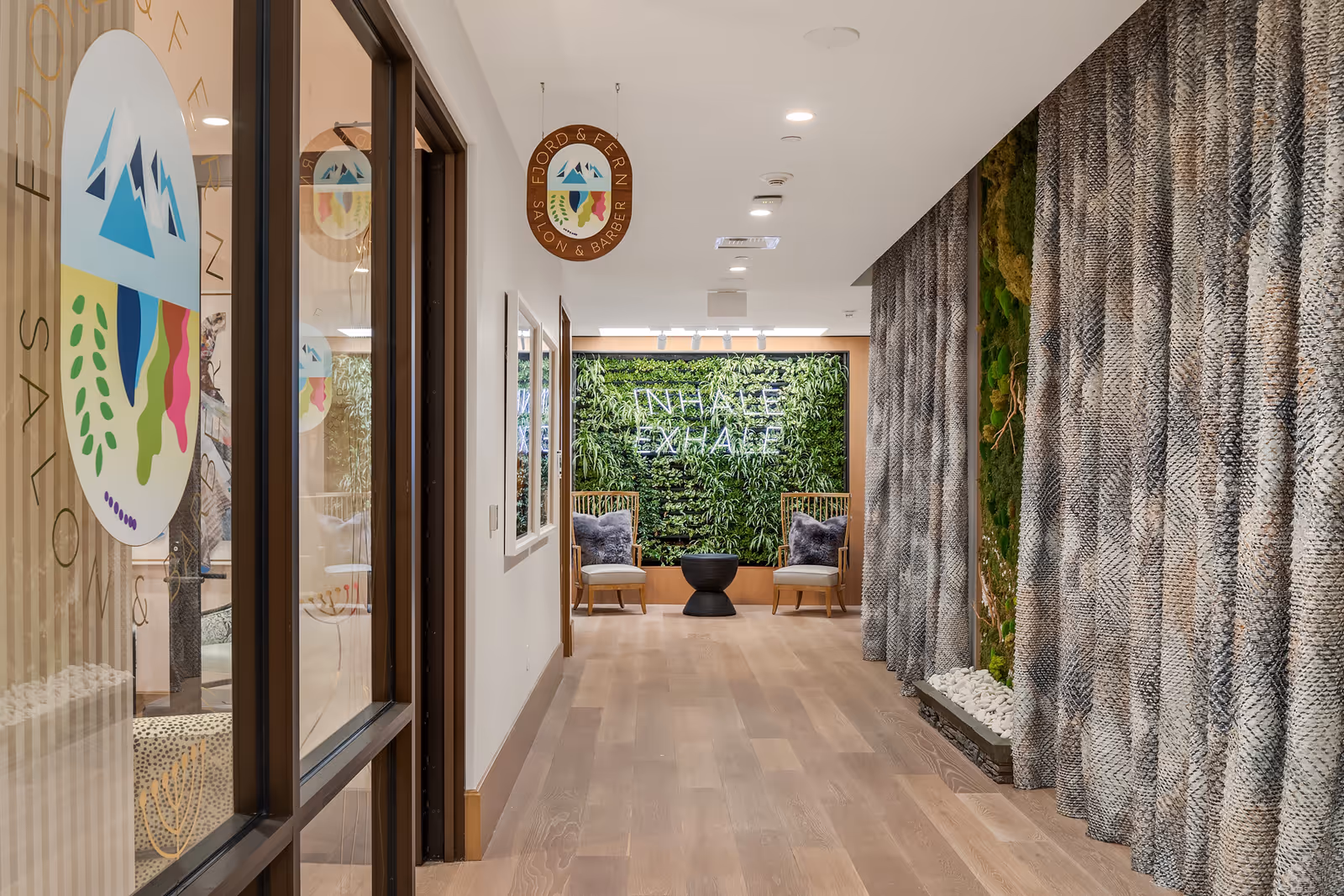A modern hallway in a senior living facility with light wood flooring, a green plant wall at the end featuring a neon sign that reads 'INHALE EXHALE', two wooden chairs with gray cushions, and a small black table between them. On the left side, there is a glass door with a colorful logo for Fjord & Fern Salon & Barber. On the right side, there is a textured curtain and a vertical garden with white stones at the base.