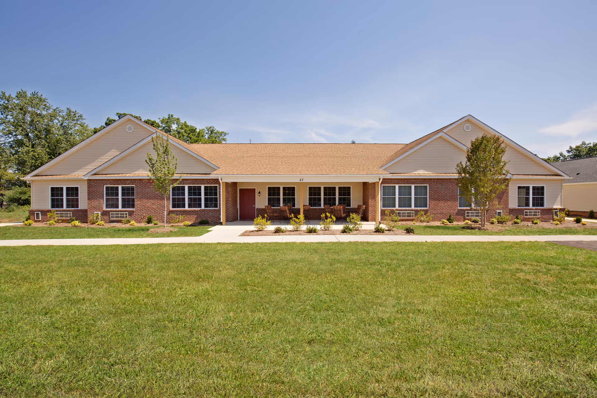 Single-story residential building with a brick and beige siding exterior, a tan shingled roof, and a covered front porch with chairs. The building is surrounded by a well-maintained lawn and small trees under a clear blue sky.