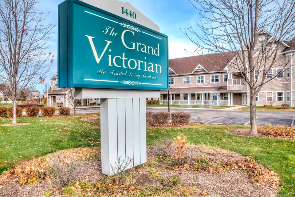 Large green sign reading "The Grand Victorian" stands in front of a multi-story assisted living building with a landscaped lawn.