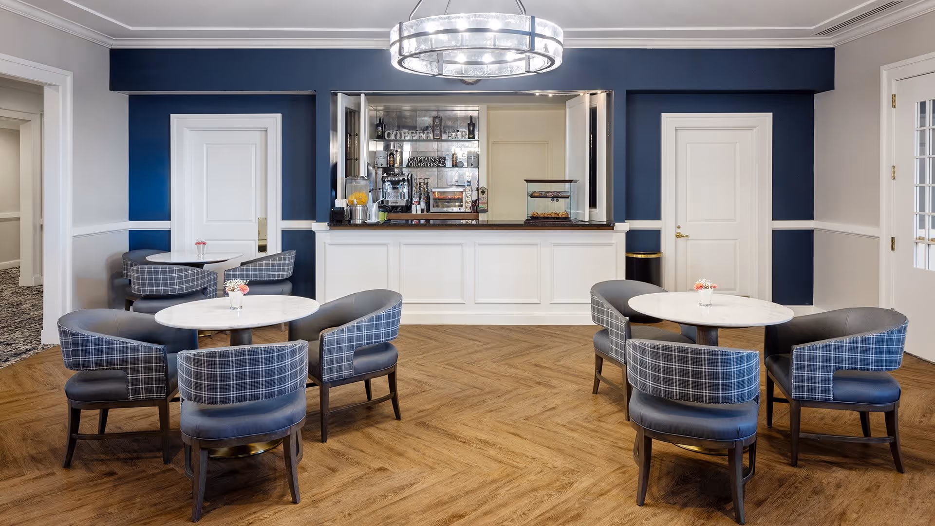 A modern dining area with round white marble tables surrounded by blue and plaid upholstered chairs. The room features a wooden floor with a herringbone pattern, blue and white walls, and a large circular chandelier hanging from the ceiling. At the back, there is a counter with a coffee bar setup including a coffee machine, juice dispenser, and pastries on display. Two white doors flank the coffee bar area.