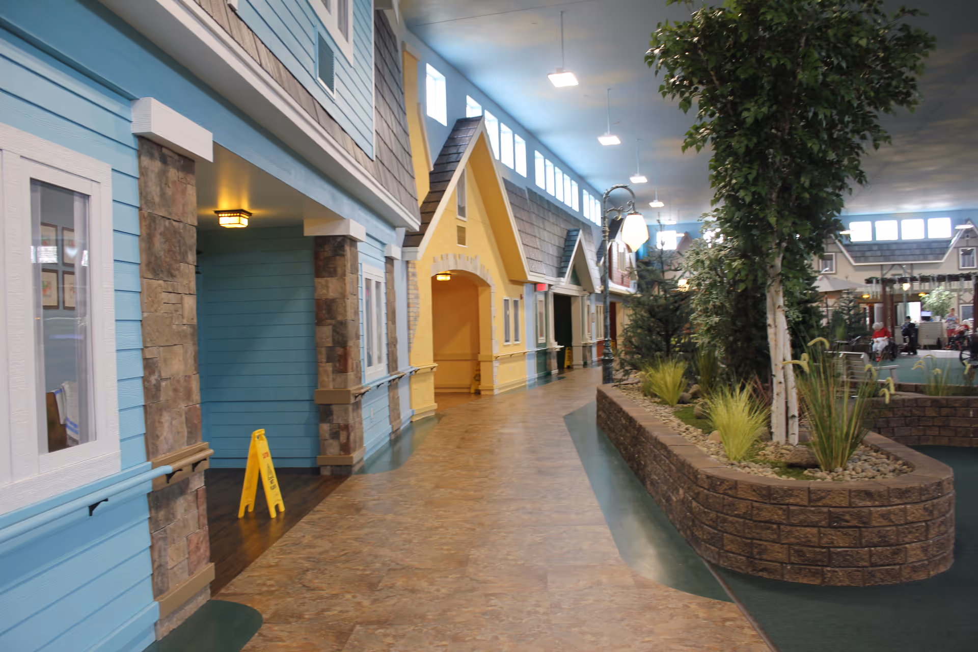 Indoor hallway designed to look like a small village street with colorful house facades, stone pillars, and a central planter with trees and plants under a high ceiling with windows and hanging lights.