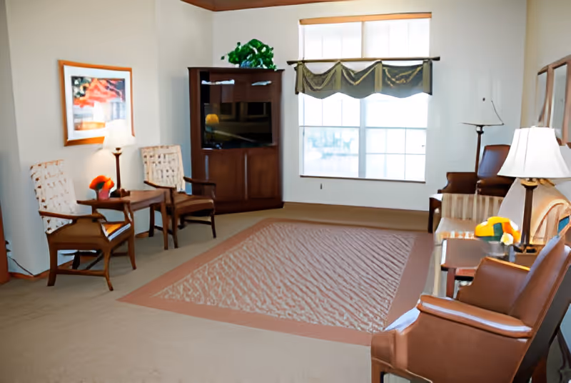 Communal living room with several upholstered chairs, side tables and lamps, a TV cabinet, and a large window with a valance.