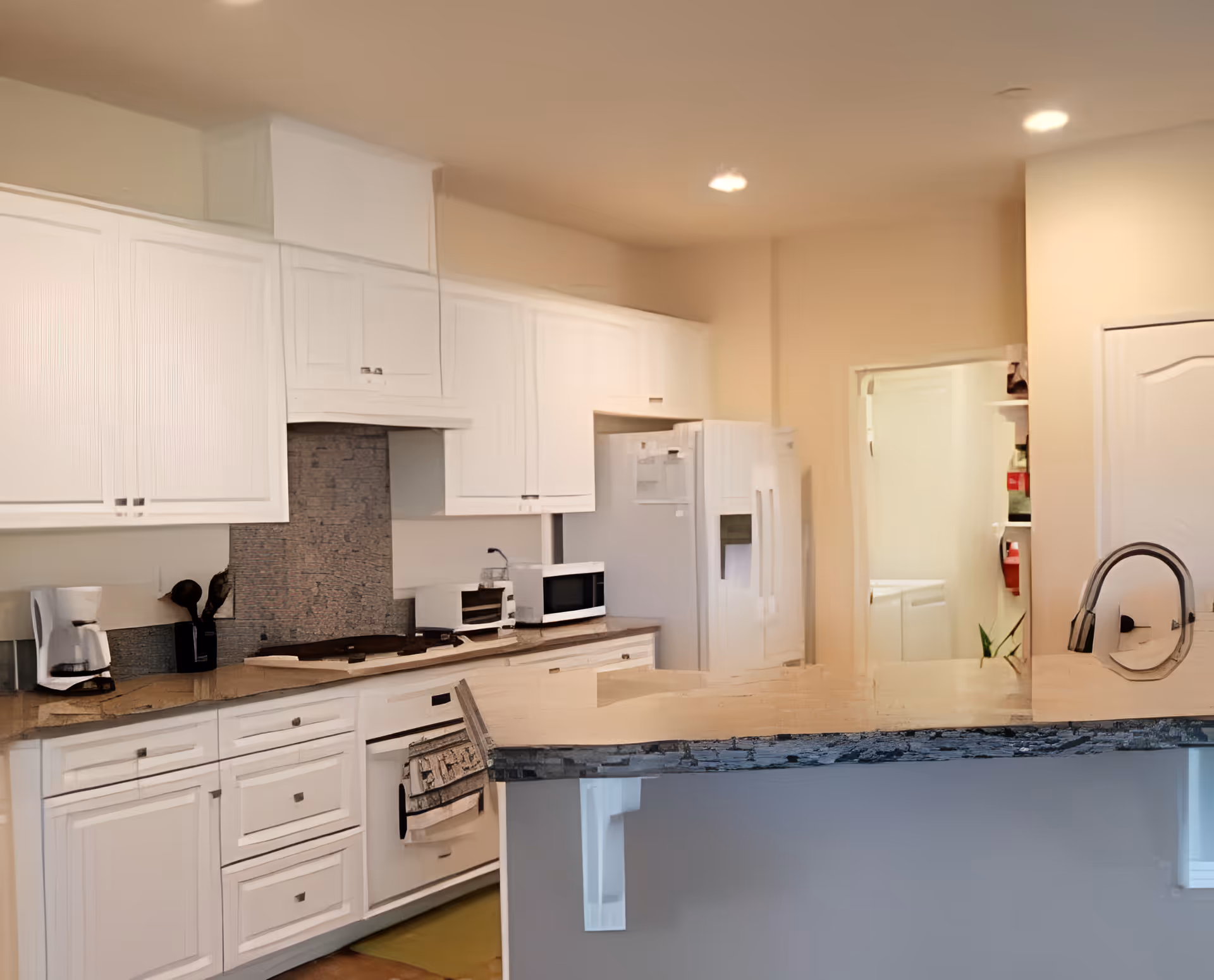 A modern kitchen with white cabinets and drawers, a built-in oven, a microwave, a coffee maker, and a refrigerator. The kitchen has a light-colored countertop with a sink and a faucet. There is a doorway leading to another room with shelves and a fire extinguisher visible.