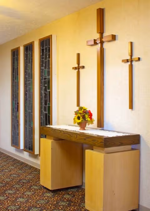 Interior hallway with three wooden crosses mounted on a beige wall above a wooden table with a white lace runner and a small vase of colorful flowers. To the left are three vertical stained glass windows. The floor is covered with a patterned carpet.