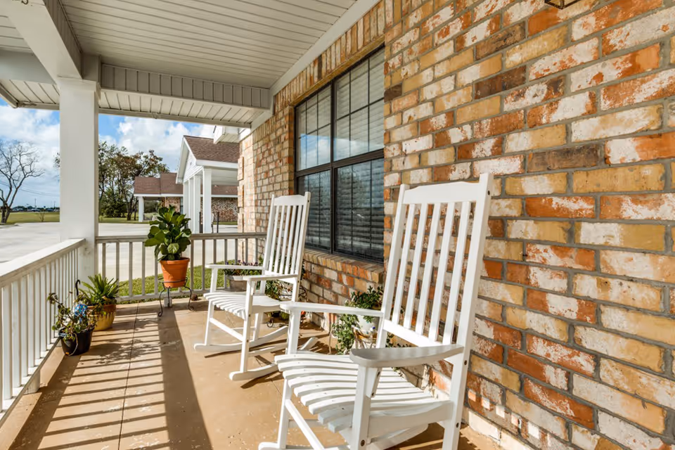 A covered porch area with two white wooden rocking chairs and several potted plants placed along the railing and near a brick wall with a large window. The porch overlooks a driveway and some trees under a partly cloudy sky.