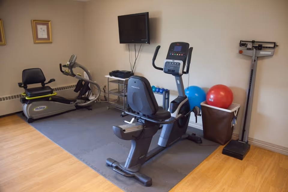 A small exercise room with two stationary recumbent bikes on a gray mat, a wall-mounted TV, a small metal cart with equipment, two exercise balls in a bin, and a standing scale against a beige wall with wooden flooring.