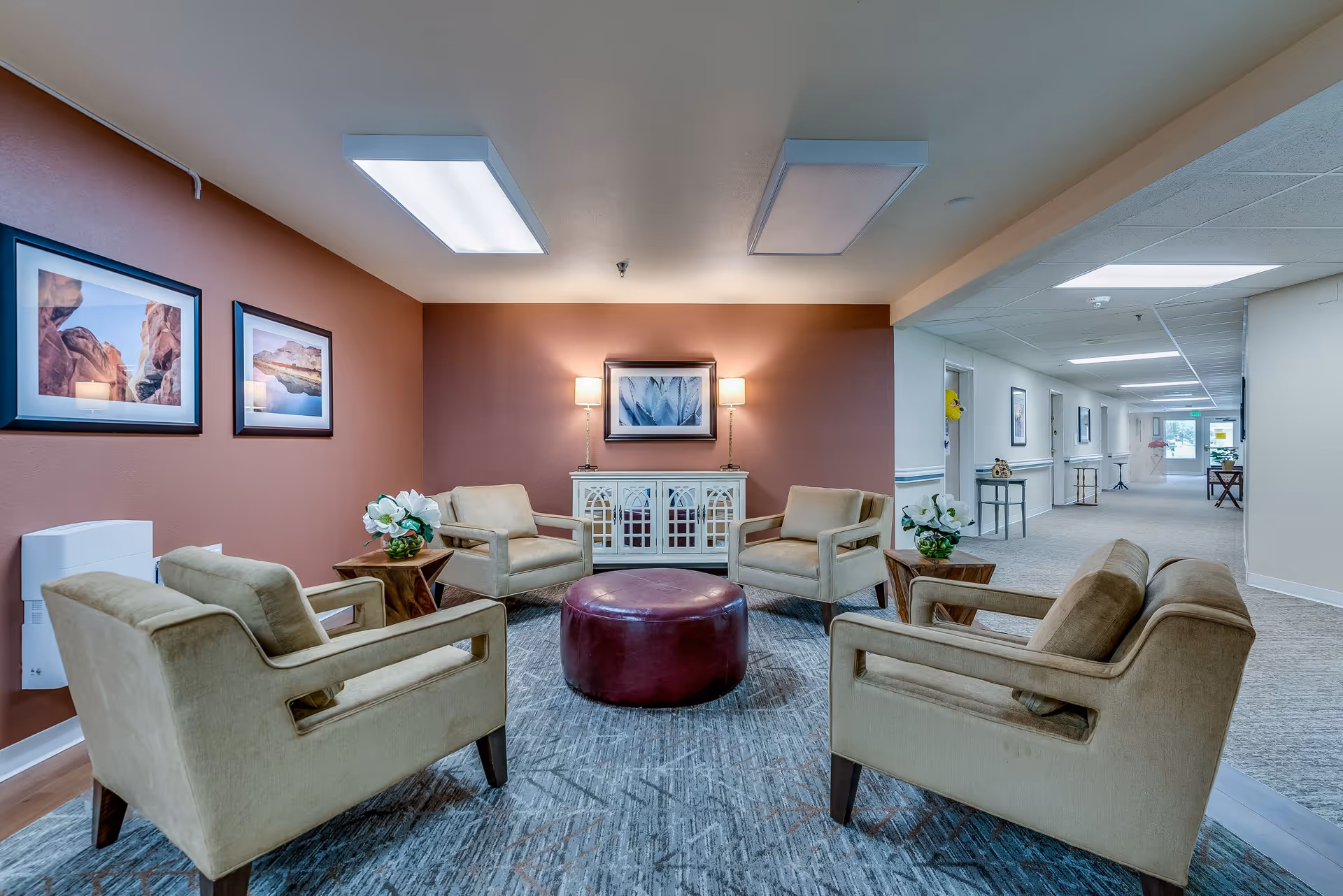 A cozy seating area in a senior living facility with four beige armchairs arranged around a round maroon ottoman on a patterned rug. The back wall is painted a warm reddish-brown and features a white cabinet with two lamps and a framed picture above it. Two framed landscape photos hang on the left wall. The seating area is adjacent to a hallway with light-colored walls and carpet, leading to a door with natural light at the end.