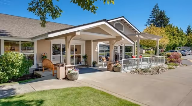 Exterior view of Crista Shores facility showing a single-story building with a covered entrance, outdoor seating area with chairs and potted plants, surrounded by greenery and a parking lot under a clear blue sky.