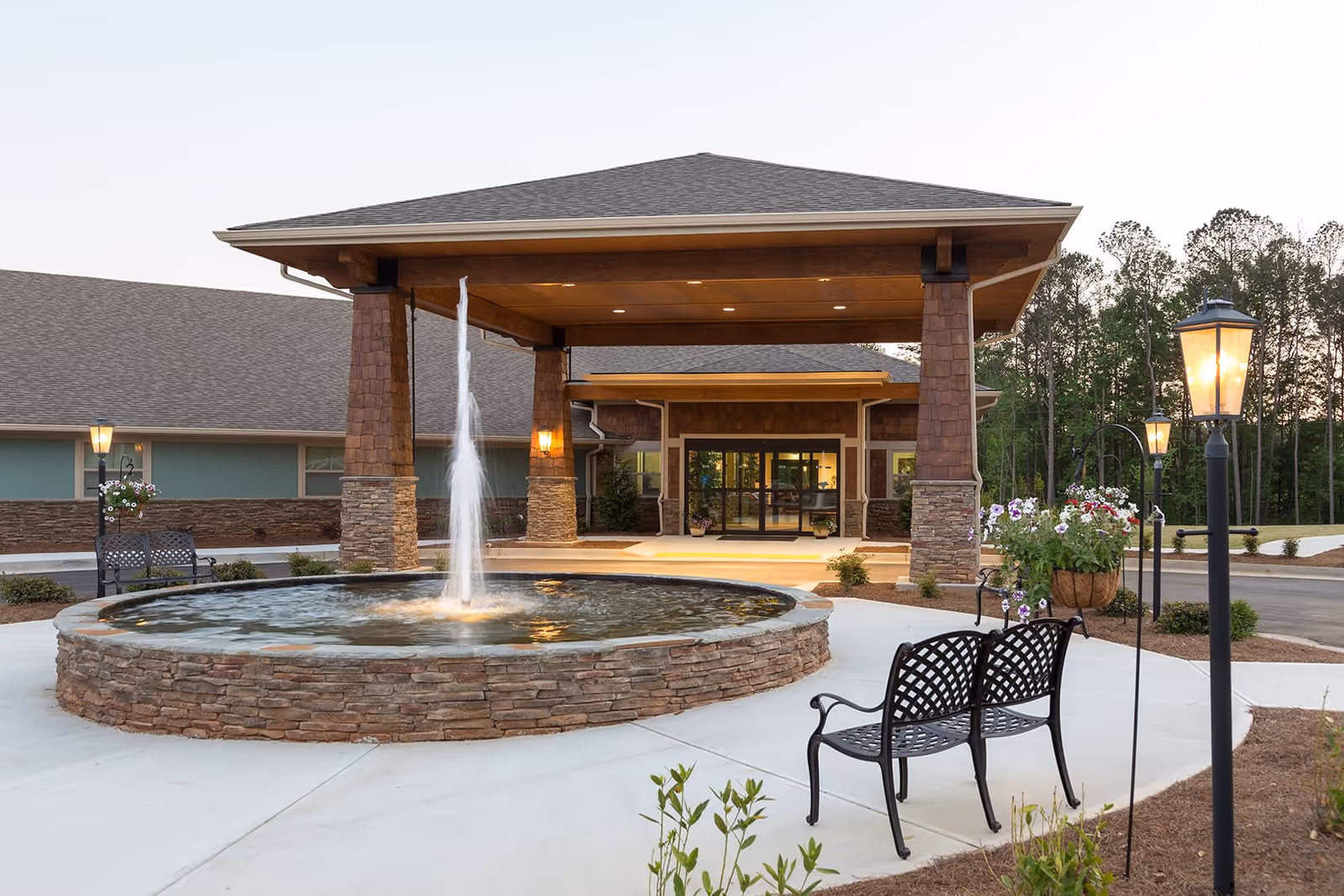 Entrance of Oaks at Hampton facility with a covered drop-off area supported by stone and wood pillars, a circular stone fountain with water jet in front, black metal benches, hanging flower baskets, and lamp posts lining the walkway surrounded by landscaped greenery.