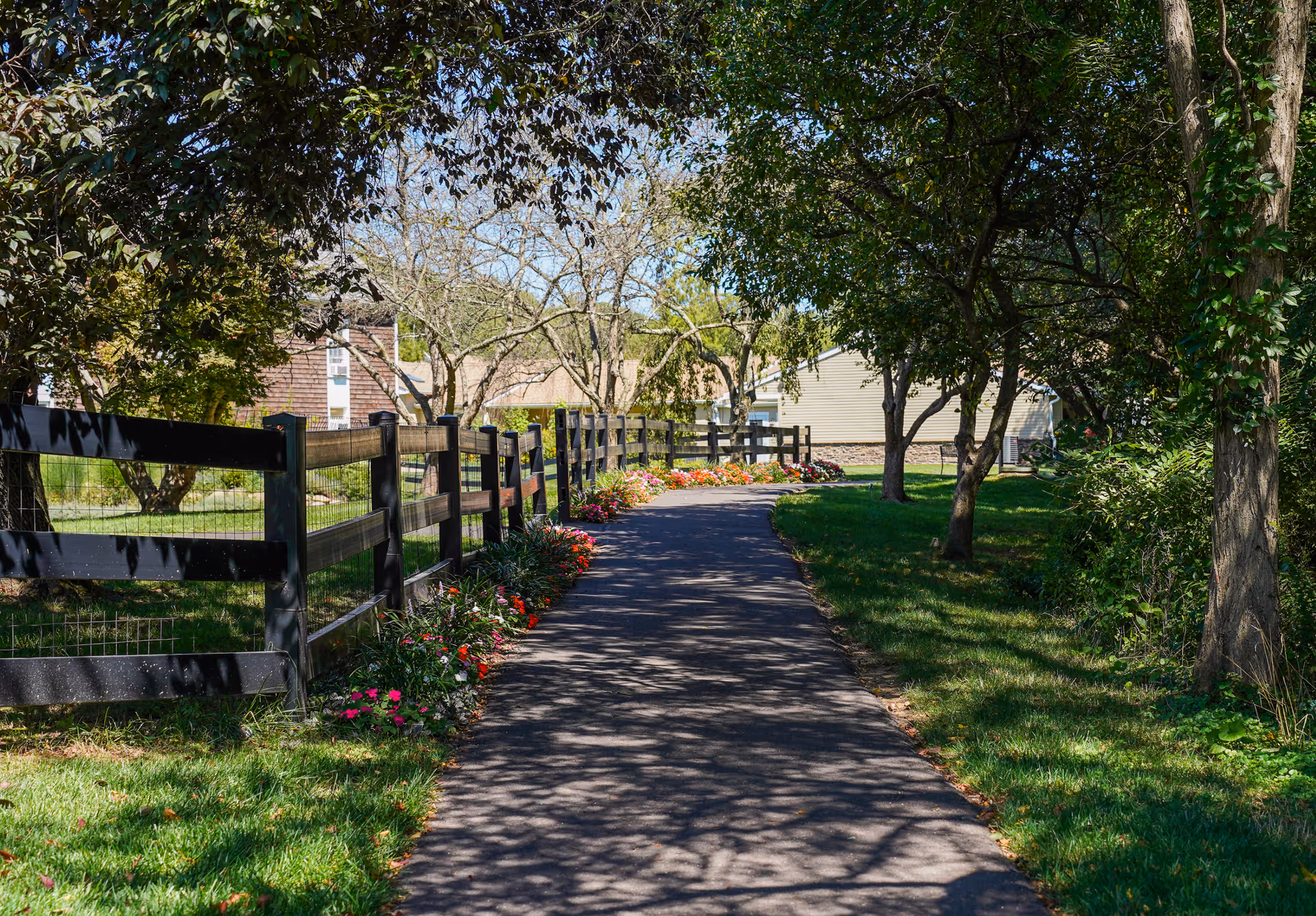 A paved walking path lined with colorful flowers on one side and a black wooden fence on the other, surrounded by green grass and trees with some buildings visible in the background under a clear blue sky.