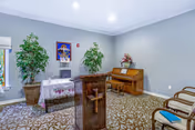 A small chapel room with a wooden lectern featuring a cross, a piano with a floral arrangement on top, two large potted plants, a religious painting on the wall, and several chairs arranged along the walls. The room has light gray walls and a patterned carpet.