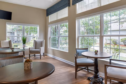 A bright and airy common area with large windows letting in natural light. The room features several round wooden tables with chairs arranged around them. There are two upholstered armchairs near a window with a small table between them holding a plant. The floor is wooden, and the walls are painted beige. Outside the windows, greenery and parts of other buildings are visible.