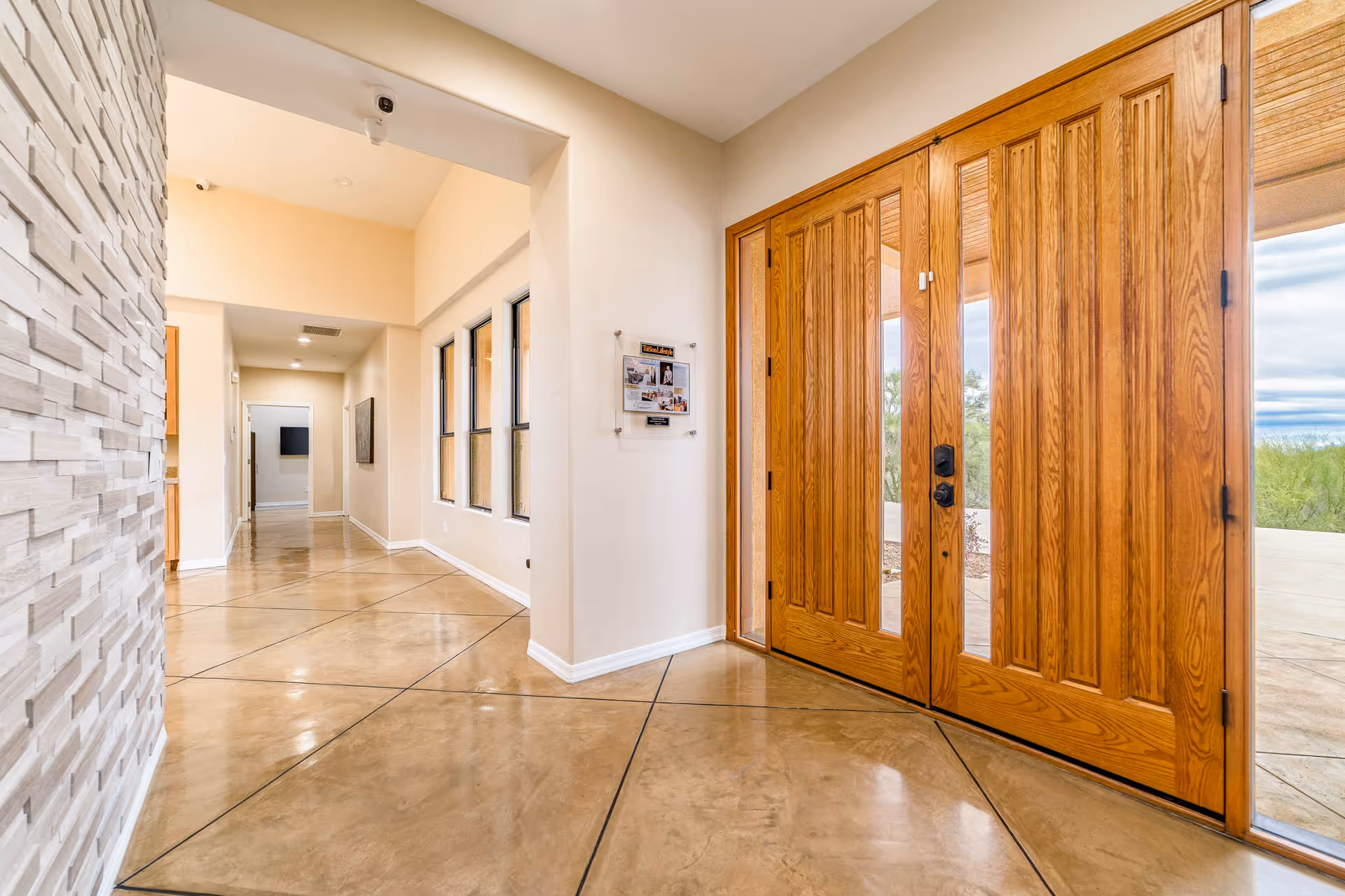 Bright foyer with large wooden double front doors, polished geometric tile floor, a textured stone wall, and a hallway with windows.